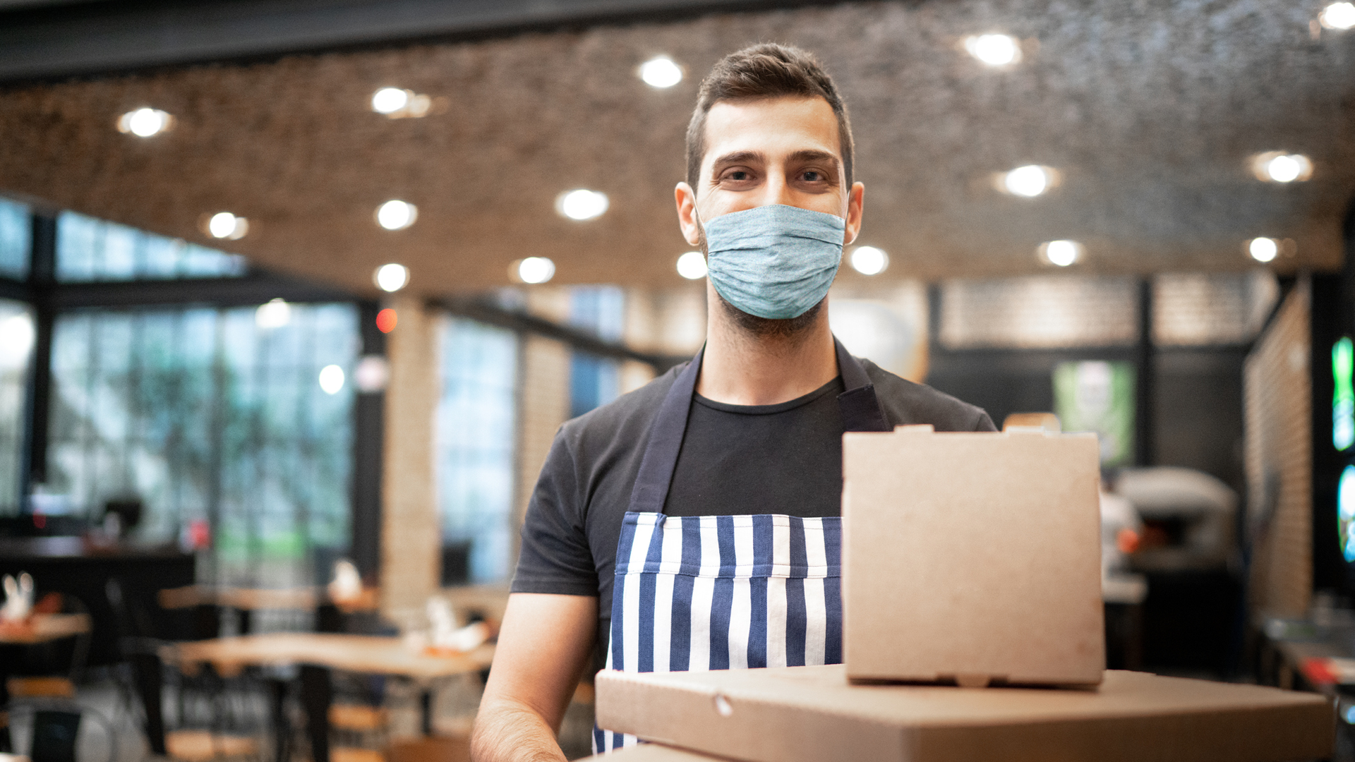 homem em restaurante, usando máscara e avental, posa em frente a uma sacola de delivery